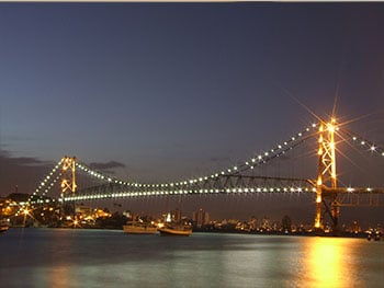 No anoitecer, vista da Ponte Hercílio Luz do lado da Ilha de Florianópolis, com suas luzes ligadas e refletidas no mar abaixo, imagem para o blog da Pousada dos Sonhos em Jurerê, Florianópolis.