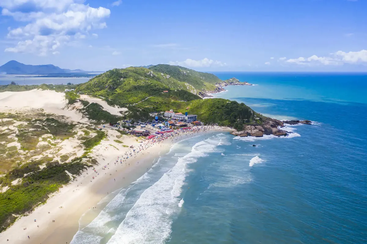 Praias de Florianópolis com mar azul, faixa de areia, dunas e vegetação ao redor.
